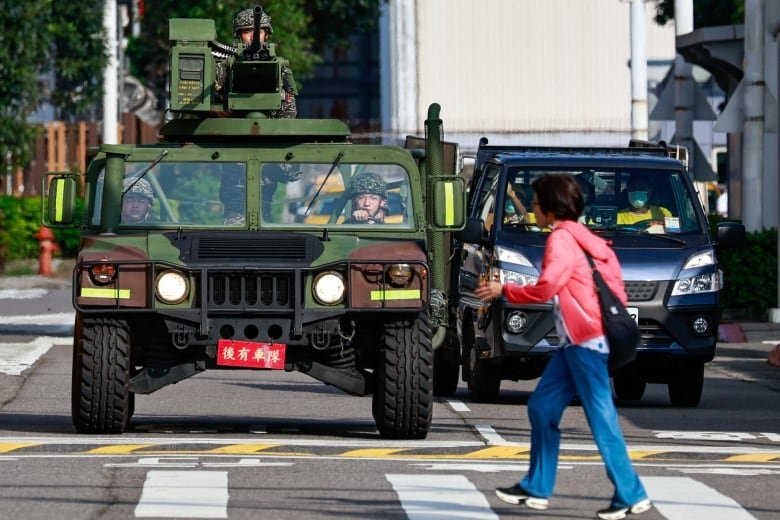 2177632177 A civilian woman walks in a cross walk in front of a stopped military vehicle topped by a manned gut turret.