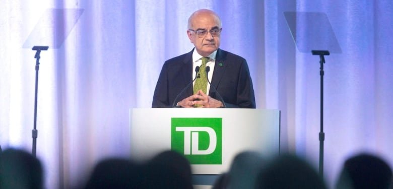 agm-td-bank-20180329 A man stands in a suit behind a podium that has the green TD logo on it. A blurry silhouette of a crowd is visible at the bottom of the image.