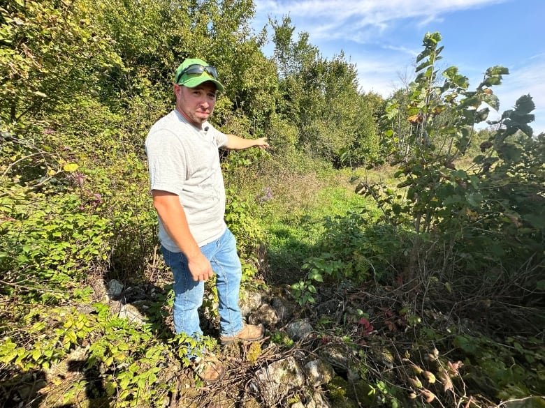 A man in a T-shirt, jeans and baseball cap points at a path created by migrants crossing through shrubbery.