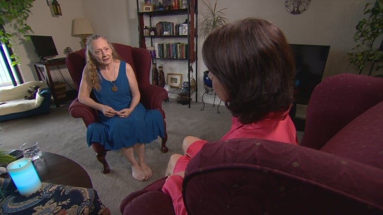 constance-mccall-and-rosa-marchitelli A woman, Constance McCall, is wearing a sleeveless blue mid-calf length dress sits in an upholstered chair with her hands in her lap, speaking to a brown-haired woman, Rosa Marchitelli, whose back is to the camera. McCall's long hair is pulled back from her face. Both women are seated in a living room with grey carpet and a bookshelf and computer monitor in the background.