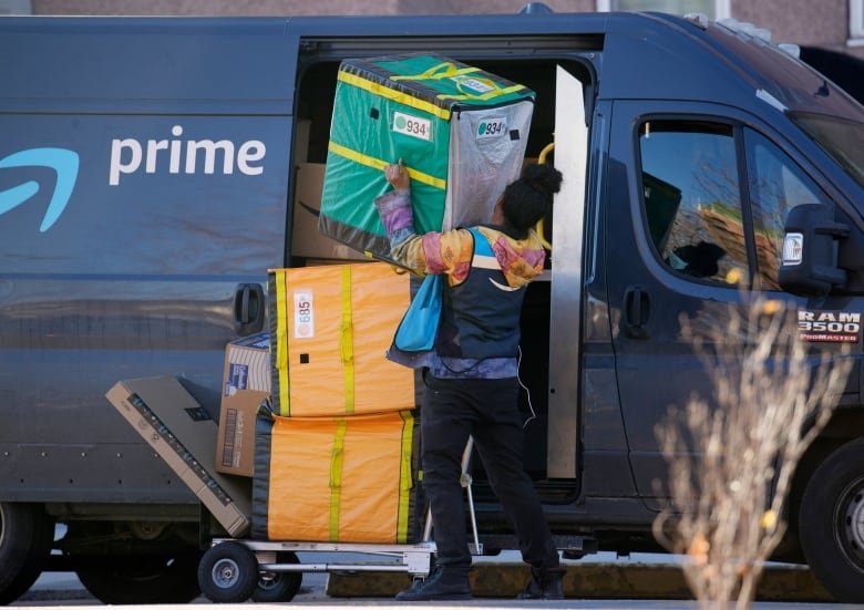 consumer-spending A worker in black-and-blue vest lifts a large package out of an Amazon delivery truck.