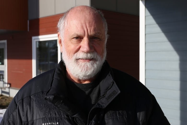 dr-louis-hugo-francescutti A smiling, mostly bald man with a white beard smiles at the camera. He is wearing a black ski jacket and stands in front of a grey and rust-coloured building.