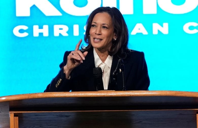 A woman with black hair, wearing a dark jacket and white blouse, gestures as she speaks into a microphone while standing at a podium.