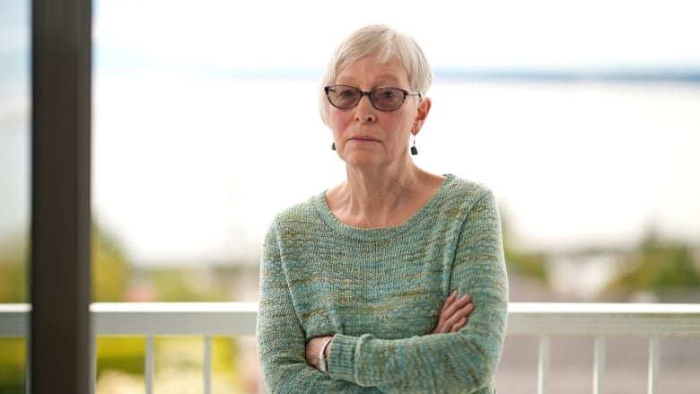 ellen-gould-2 An older white woman wearing darkened glasses and a green and blue knit sweater stands on her balcony with her arms crossed.
