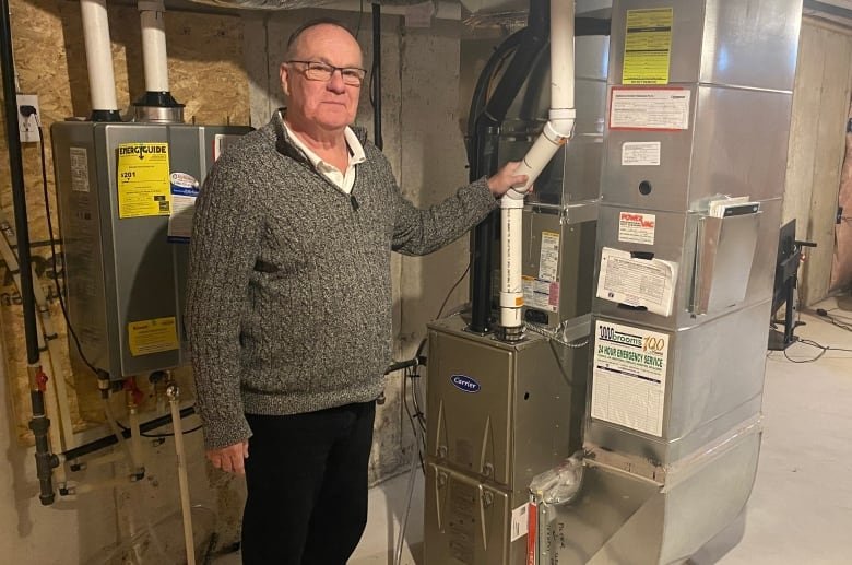 An older man with glasses wearing a grey sweater and dark pants stands in a basement with his hand on a furnace pipe.