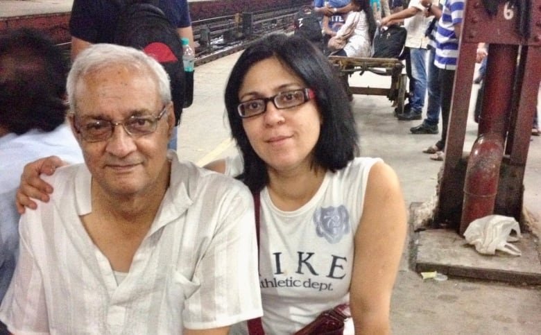 harish-and-shanu An aged south Asian man and a south Asian woman sit on a bench on a busy train platform. The woman has her right arm around the man.