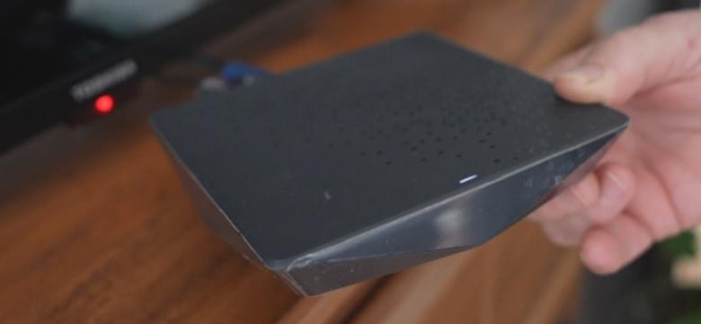 ignite-tv-box A hand holds a small, black gadget which is attached by cable to a TV.