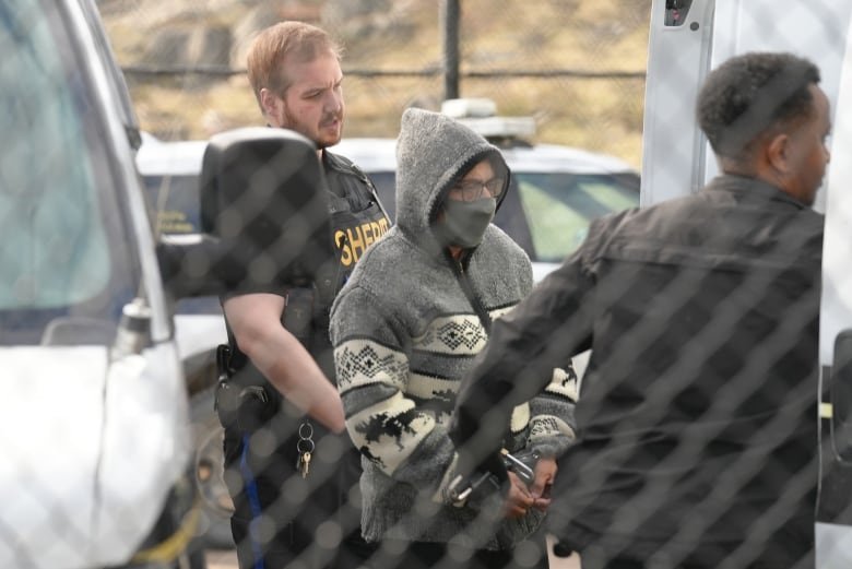 karima-manji A woman wearing a grey sweater with the hood up and a grey face mask and hand cuffs is seen through a fence with two sheriffs on either side of her.