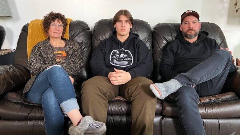 kelly-left-wil-centre-and-kurt-krotenko Three people sit on a brown leather couch wearing neutral facial expressions. On the left is a cross-legged woman with short brown curly hair and glasses. She is wearing jeans, a dark cardigan sweater over a grey and orange t shirt, and grey beaded moccasin style slippers. In the centre is a young man with short brown straight hair sitting straight with hands folded in his lap, brown pants, and a black hoodie sweatshirt. On the left sits an older man with one leg crossed at the knee. He has a black and grey beard, dark baseball style hat, grey jeans, and grey wool socks.