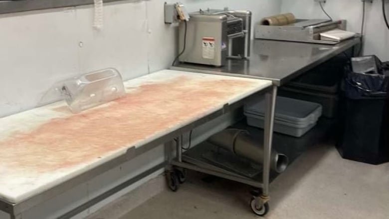 meat-department-room-at-gateway-co-op-store-canora-sask In the foreground is a white table with blood on it, next to a wheeled stainless steel cart with a meat wrapping machine and other equipment. On a lower shelf of the cart are some stacked white plastic bins. There is a garbage bin with a black plastic bag in it to the right of the meat wrapper.