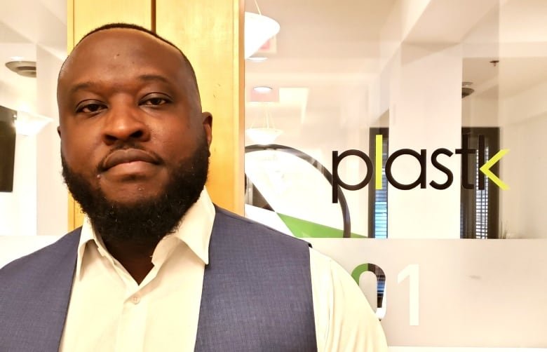 motola-omobamiduro A bearded man wearing a white collared shirt and blue vest faces the camera with a slight smile. He is standing in from of a pair of glass doors printed with the Plastk logo in green and black text.