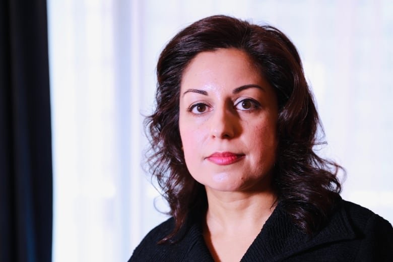 nafissa-ismail A woman with dark brown hair and a black shirt sitting in front of white curtain and looking directly at the camera.