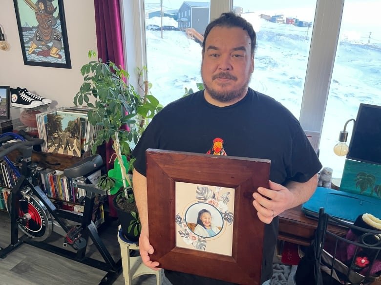 noah-noah A man stands in a home holding up a framed photo of an elderly woman.