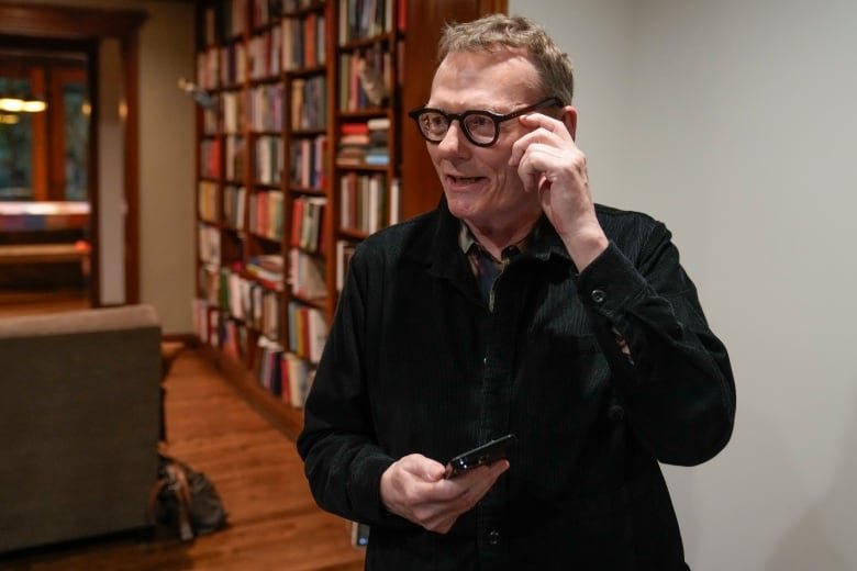 A man wearing glasses holds his smartphone as he stands near a bookcase.