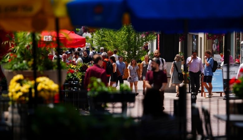 ottawa-hot-weather-20240618 The photo is taken through the shadow of a bunch of umbrellas, which are silhouetted hazily along the top of the image. The focus is crowds of people walking down the street beyond the umbrellas. It is very sunny out.