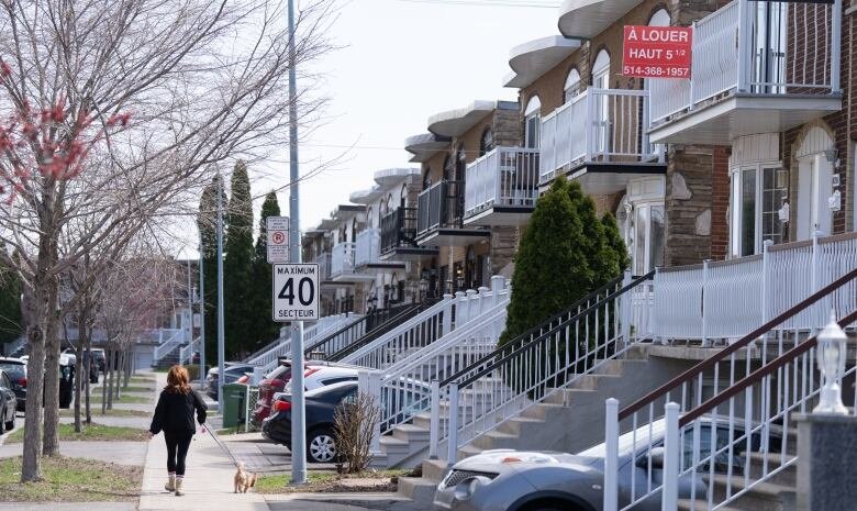 que-homes-rent-20240423 A residential street is shown, with a person walking down the sidewalk. Trees are bare of leaves. A red sign hangs outside of the second floor of a house, attached to the balconey.