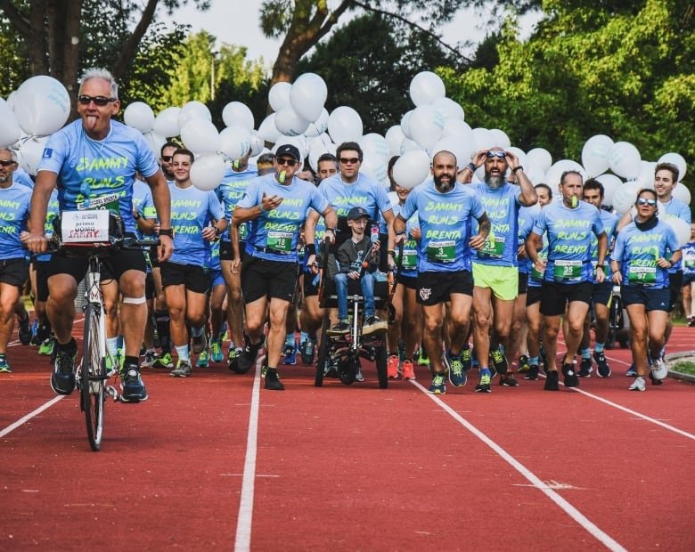 A group of people run on a track, carrying white balloons, and wearing in matching sky-blue shirts that read "Sammy's Runners." On the left, a man in the front leads the group on a bicycle and sticks out his tongue. At the centre of the group is a small man in a wheelchair.