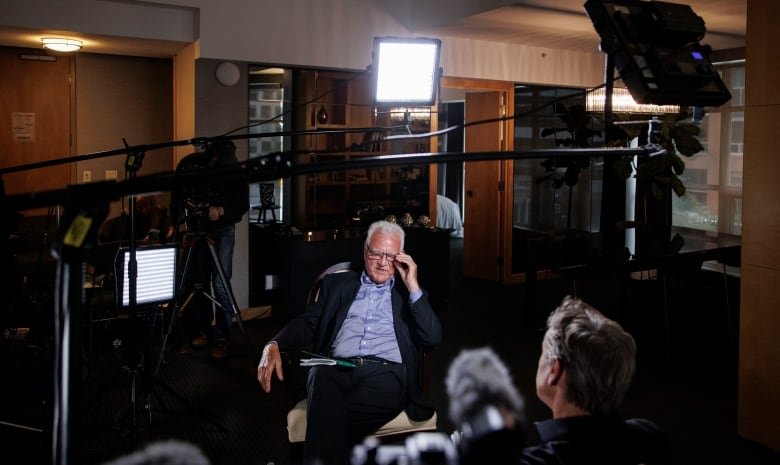 A man in a suit sits in the middle of a large, darkened room, surrounded by lights and TV cameras.