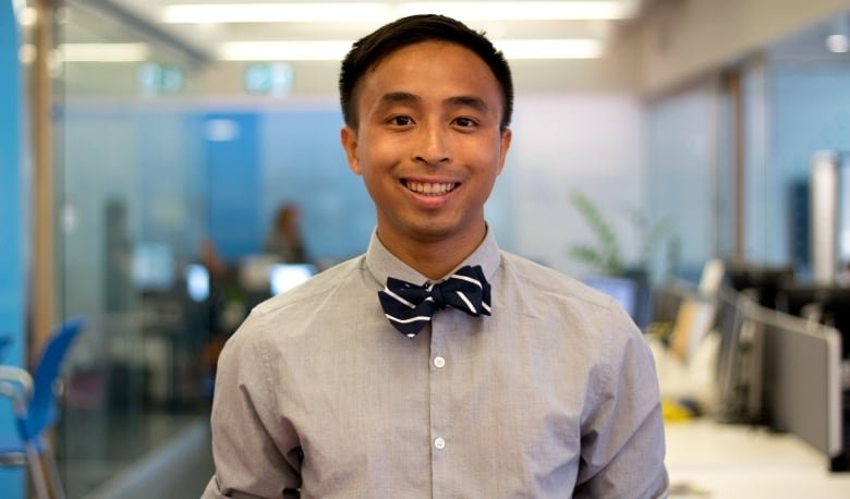 viet-vu An Asian man with short hair wearing a grey shirt with a black striped bow tie stands in the middle of an open concept office.