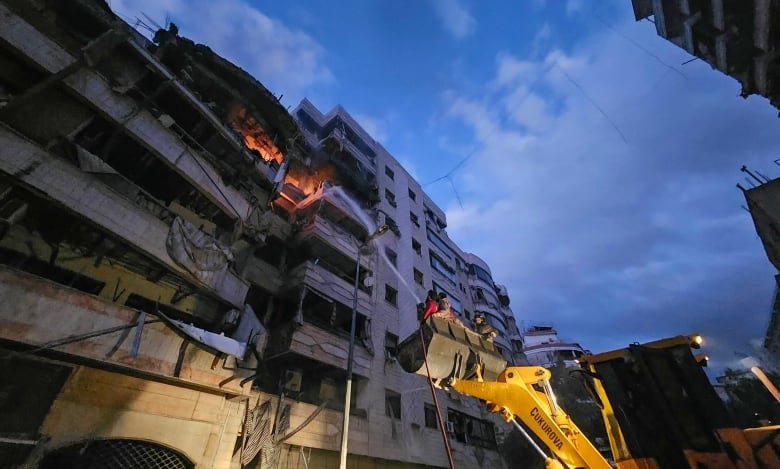 Firefighters standing on the raised plough of a bulldozer use a fire hose to spray water at a burning apartment building.