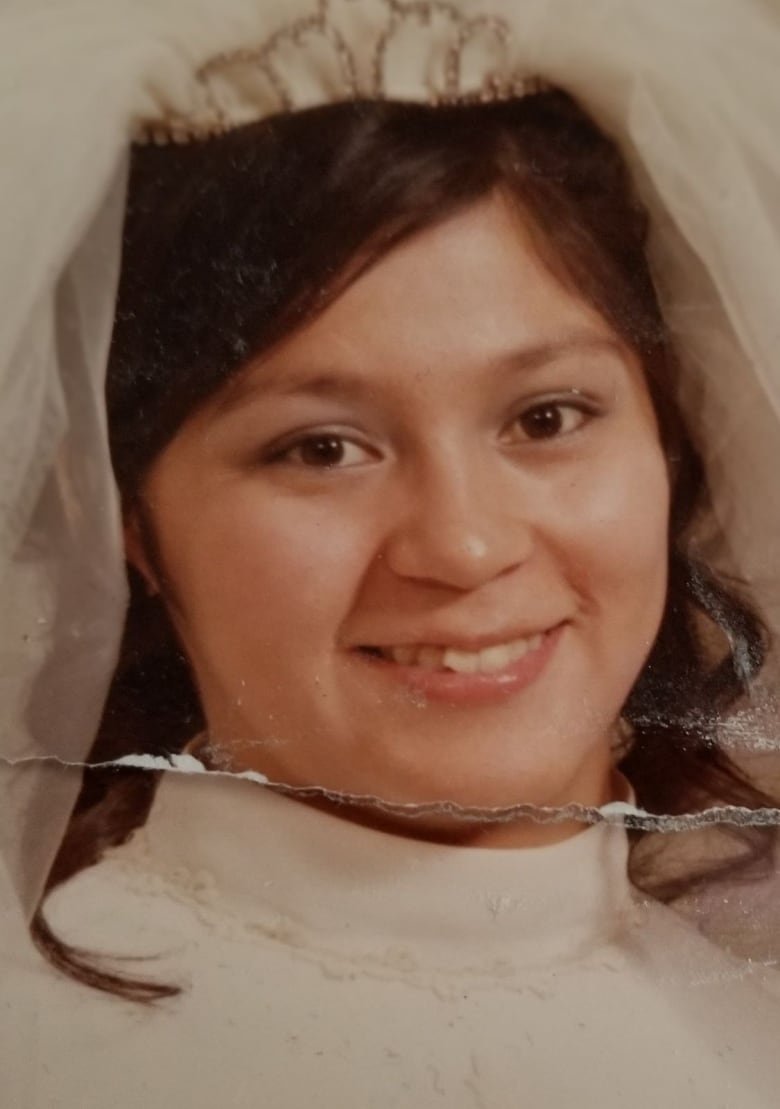 An old photograph of a young woman on her wedding day. She is smiling, wearing a white dress and a small vail. There is a rip in the photo.