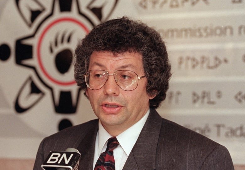 A man speaks into a microphone with a banner in the background behind him.