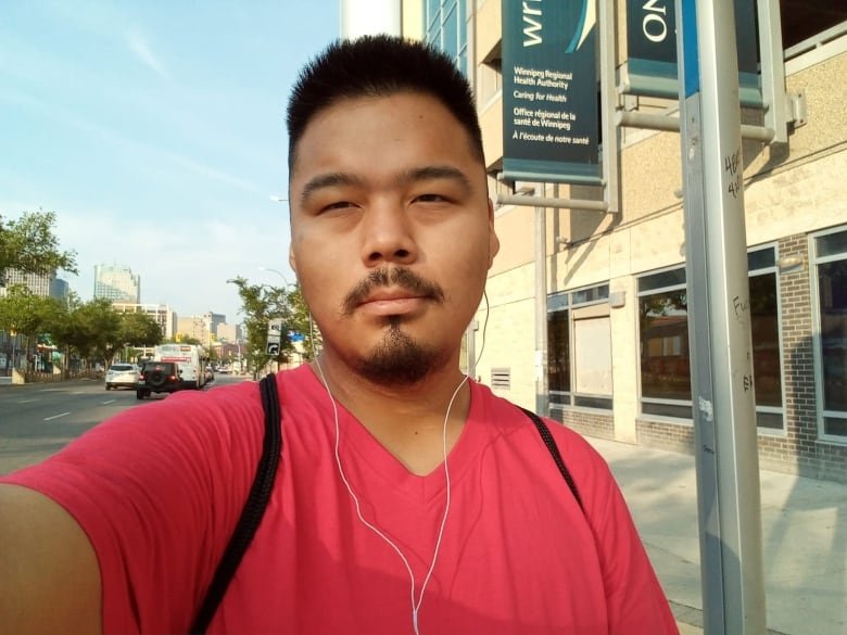 A man with short, spiked black hair and a goatee stands outside in a red T-shirt. He has earbuds on and the white wire is seen leading from the ears to the bottom of the photo. A streetscape is seen in the background.