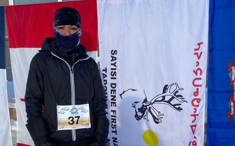 A woman wears all black clothing with a balaclava outside in front of multiple country flags.