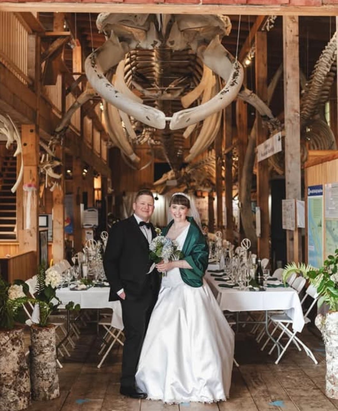emily-gatto a man in a tux stands beside a woman in a white wedding dress, underneath a suspended whale skeleton