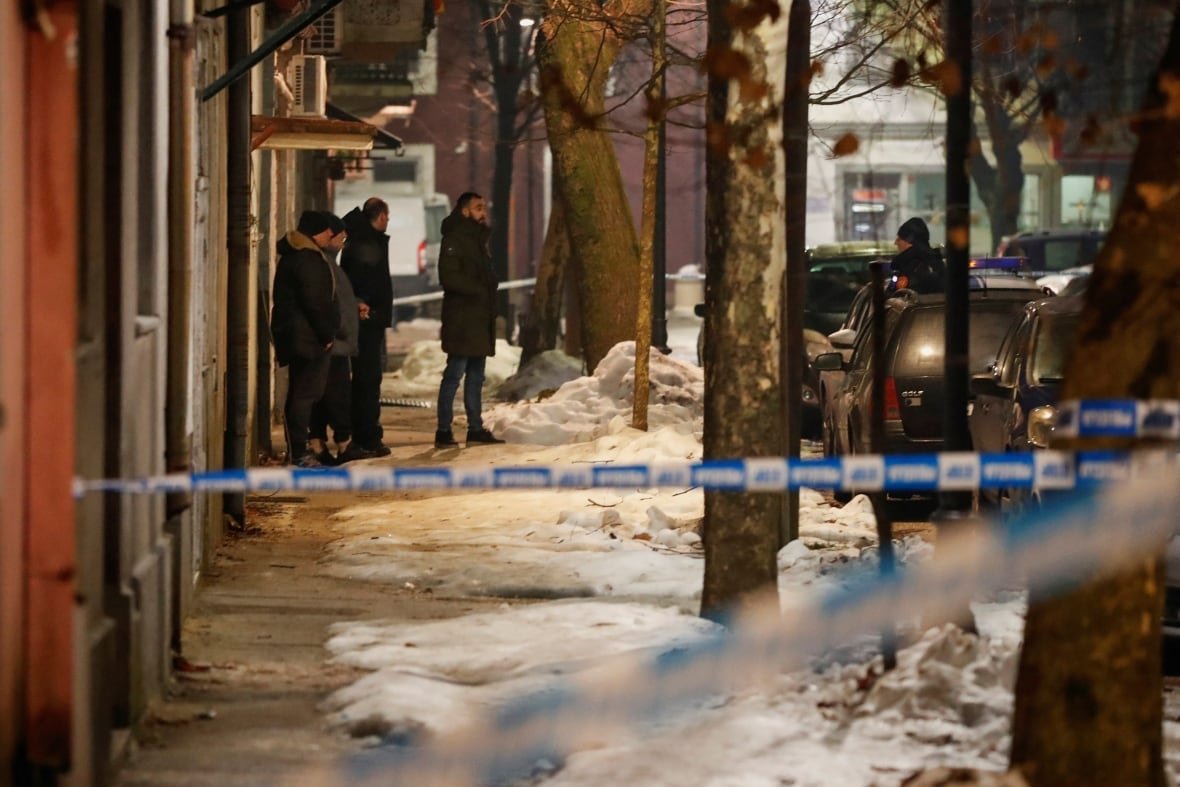 montenegro-shooting Part of a sidewalk is cordoned off by police tape as people gather behind it.
