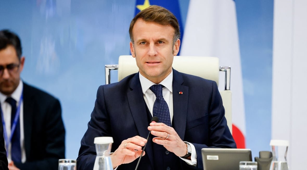 A man in a suit is in the centre of the image, with another man visible at the left side of the image. The man in the centre appears to be in the middle of speaking or about to speak and has both hands slightly raised near the microphone on the desk he is sitting at.