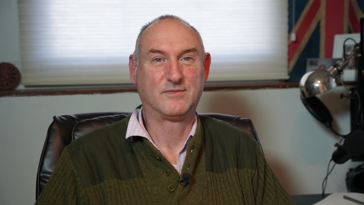 giles-crouch A balding man with close-cropped grey hair wears a green sweater and sits in an office.