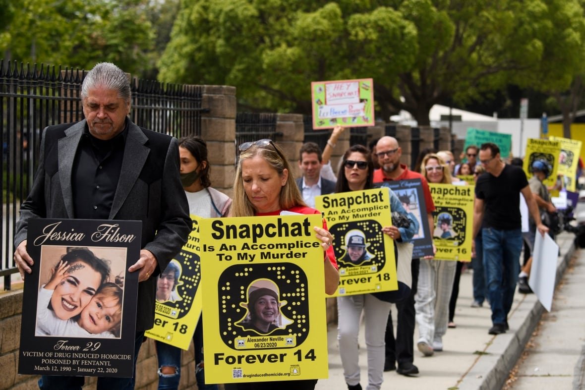 snapchat-protest A group of people walk along a sidewalk carrying yellow placards reading 'Snapchat is an accomplice to my murder' with the Snapchat logo and pictures of their loved ones and the age at which they died of drug overdoses.