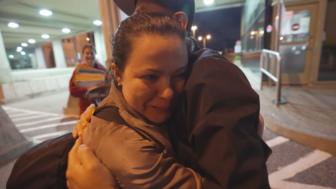 aracely-serrano-and-israel-serrano A woman hugs a man with a baseball cap. A woman stands in the background looking at them. They are at the Canadian port of entry in Niagara Falls, Ontario.