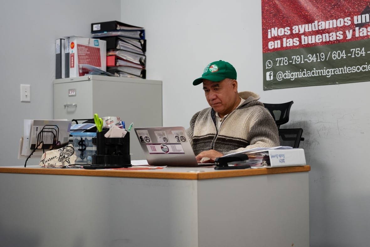 migrant-rights A man in a green baseball cap sits and a desk working on a laptop computer.