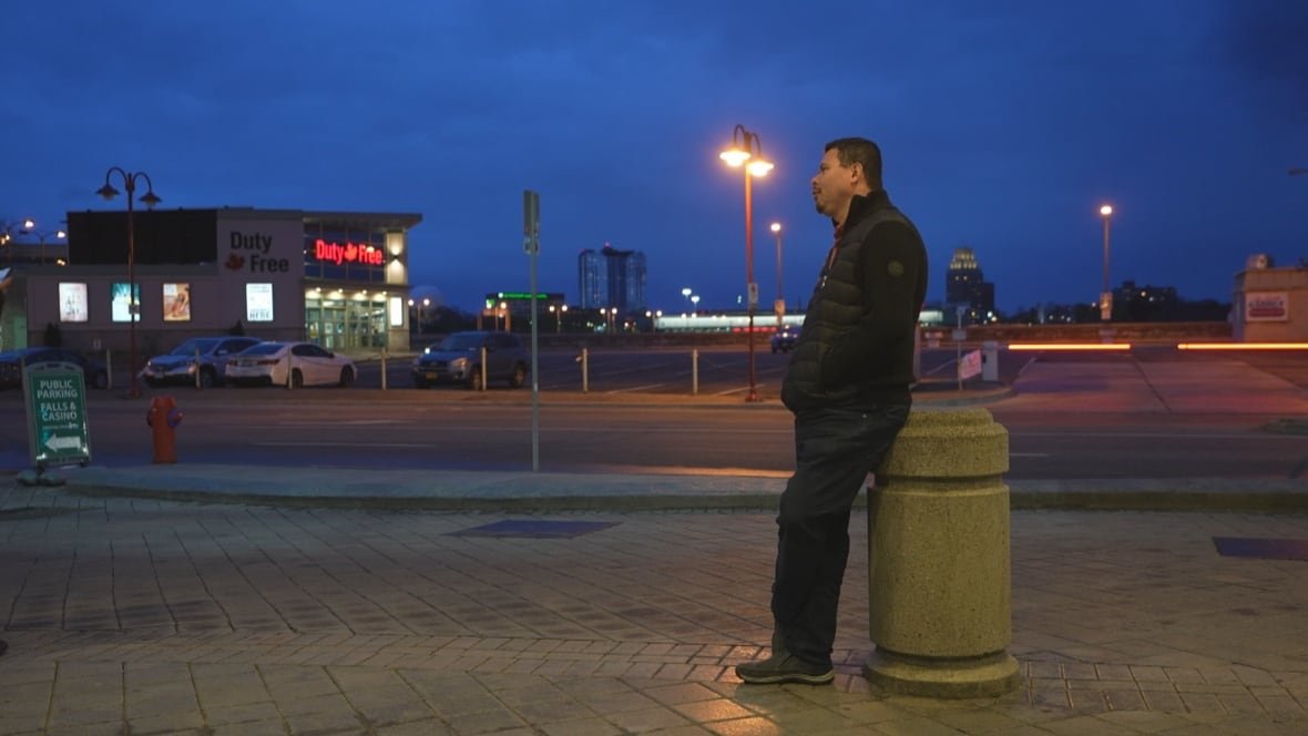 miguel-serrano A man stands on sidewalk looking to the left while leaning on a short concrete post.