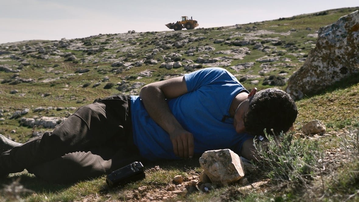 no-other-land A man lies on the ground on a grassy hill studded with rocks. A bulldozer is visible at the top of the hill beyond him.
