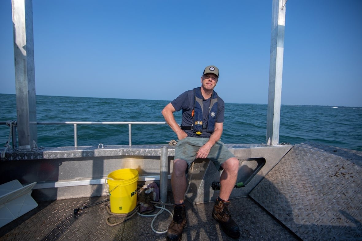 aaron-fisk-lake-erie A man wearing a blue cap, shorts and a t-shirt sits on the edge of a small metal research boat. His hand is on his hip and he's slightly grinning. Behind him, blue water meets the horizon. A yellow bucket, rope and various gear sits at his feet. The sun is shining.