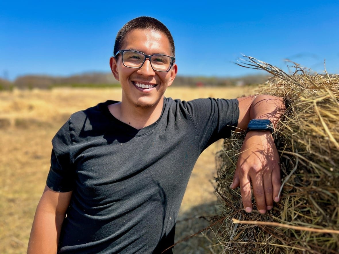buffalo-ranch-manager-jeremy-robert-chartrand-kayseas A young man is standing on the bison pasture, leaning against a haybale with a smile
