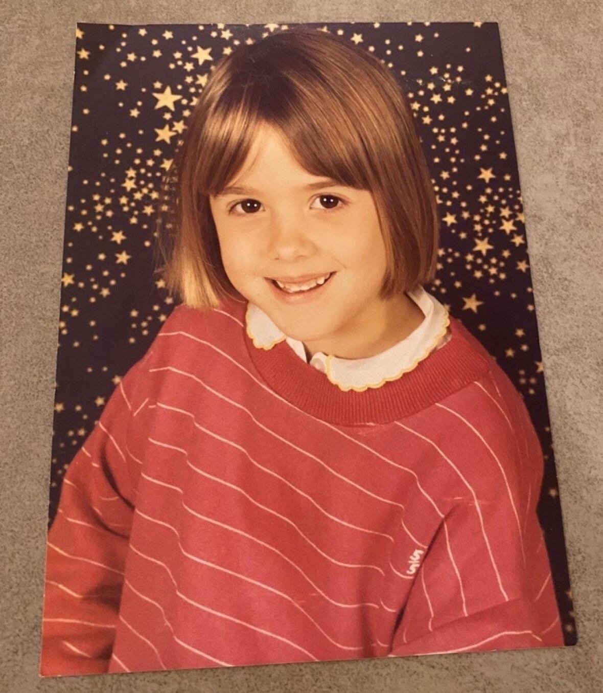 caroline-darian-childhood-photo A studio portrait of a young girl with brown hair wearing a red-and-white striped sweater.