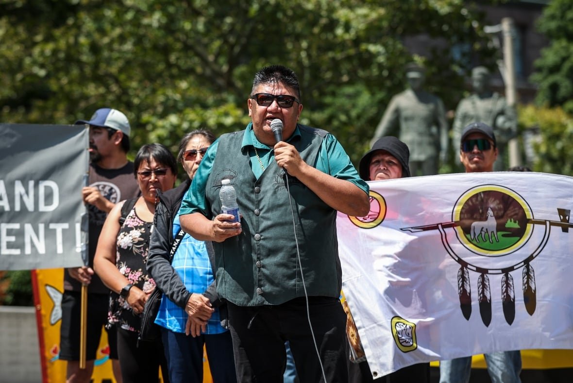 chief-chris-moonias A person stands and speaks into a microphone. A crowd of people stand behind him, holding a large flag.