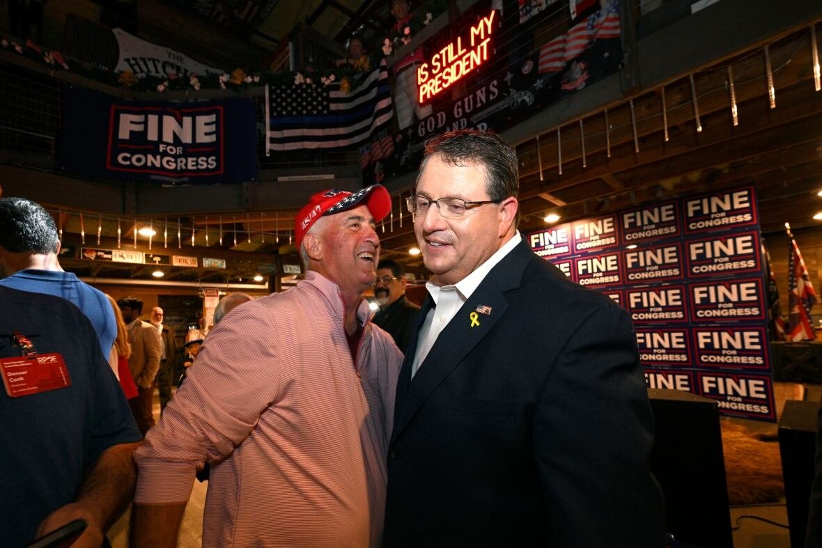 election-2025-florida-special-election A bespectacled man in a blazer smiles while in close proximity to an older man wearing a baseball cap.