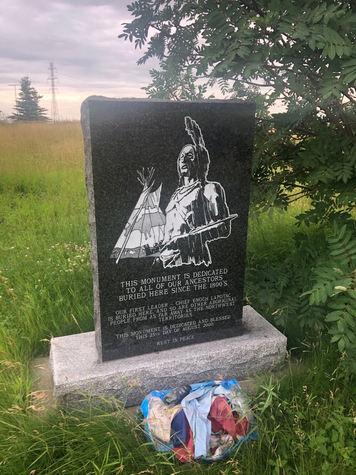 enoch-cemetery-1 A black stone, which has an etching of a tepee and a Cree man in traditional garb, in a grassy field.