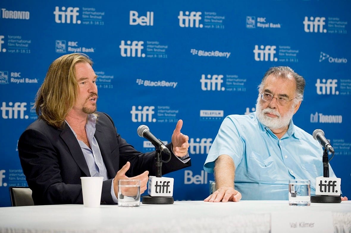 film-tiff-twixt-20110912 A blonde haired man with a goattee speaks at a long table while next to him a bespectacled older man with a grey beard listens.