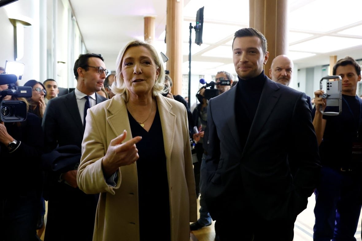 france-politics A blonde haired woman with hair to her shoulders wearing a light colored jacket and a younger, dark haired man with a beard wearing a turtleneck are shown.