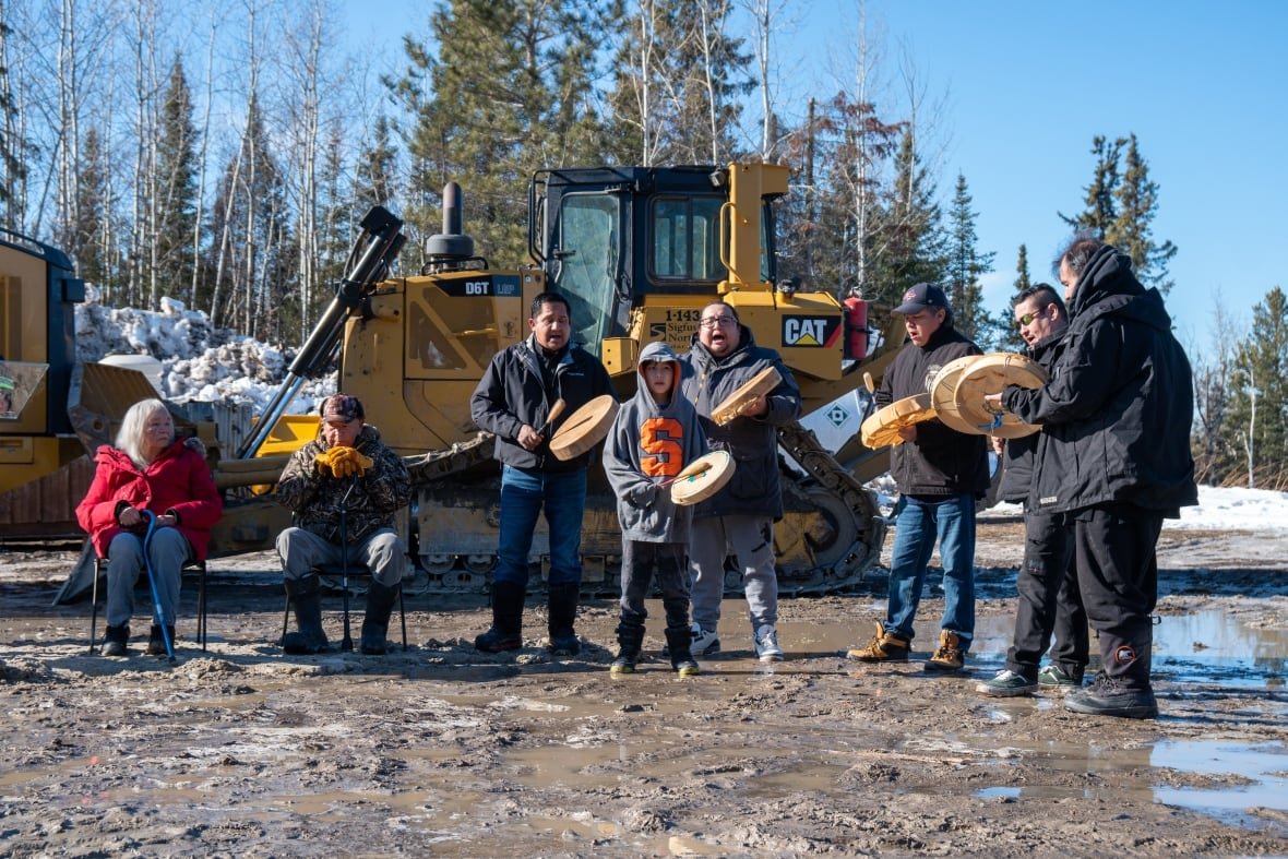 grassy-narrows-groundbreaking-1 A group of people are seen using hand drums at a muddy construction site.