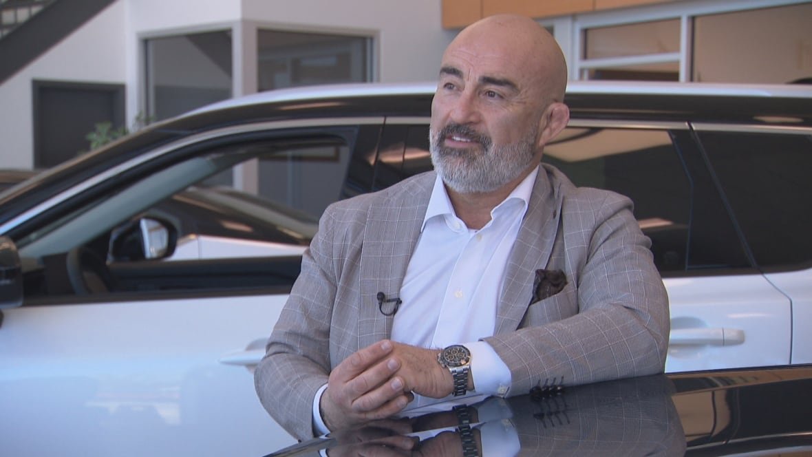 A bald man with a beard and a suit stands with his arm on a car, looking off into the distance