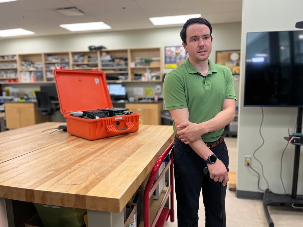 jacob-woods A man in a green shirt stands in front of a table with an open box containing communications equipment in it.