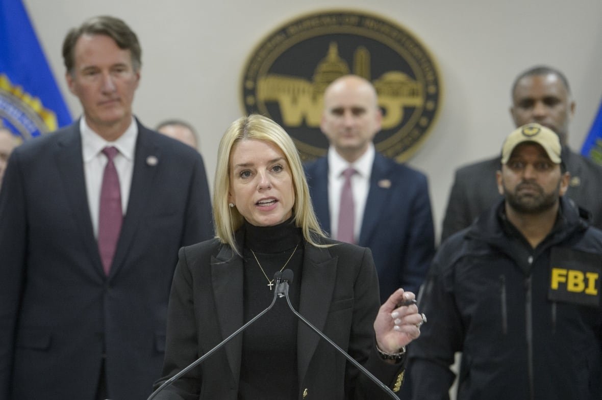 justice-department-gang-leader A blond-haired woman wearing a dark blazer and dark turtleneck speaks at a podium, with other officials, mostly men, shown in the background.