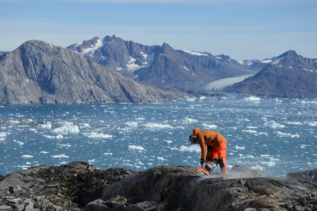 lev-tarasov Using a saw, a man cuts into rock in Greenland with tall mountains and icebergs dotting the water below.